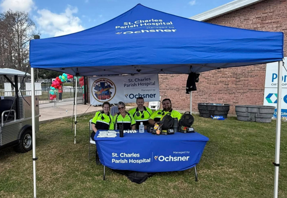 Four emergency medical staff sit at a table under a St. Charles Parish Hospital tent, with medical equipment displayed, at an outdoor event.