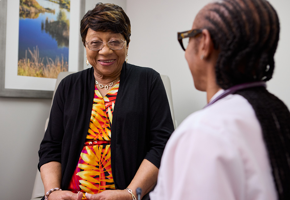 An older woman in a colorful dress sits and smiles while talking to a healthcare professional