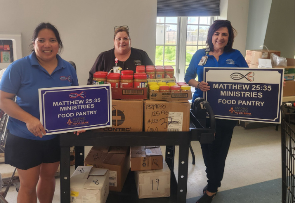 Three women stand behind a cart with food supplies and boxes, holding "Matthew 25:35 Ministries Food Pantry" signs in a room with a window.