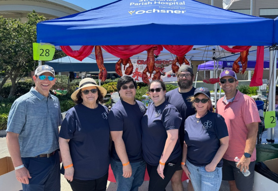 Seven people stand in front of a blue Ochsner tent at an outdoor event, posing for a group photo. Red decorations and numbered signs are visible in the background.