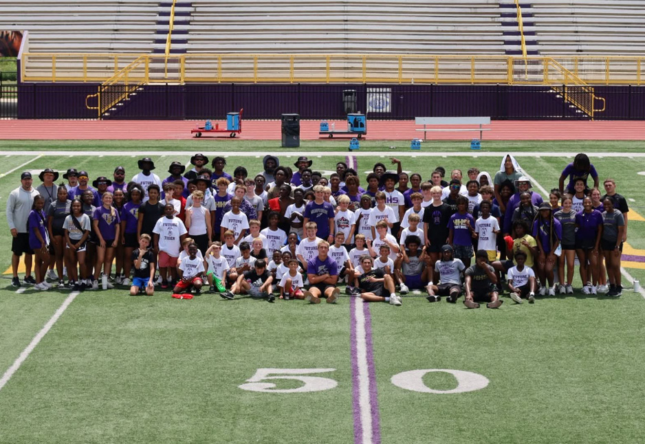 A large group of students and coaches pose for a photo on a football field, centered on the 50-yard line, with empty stadium bleachers in the background.