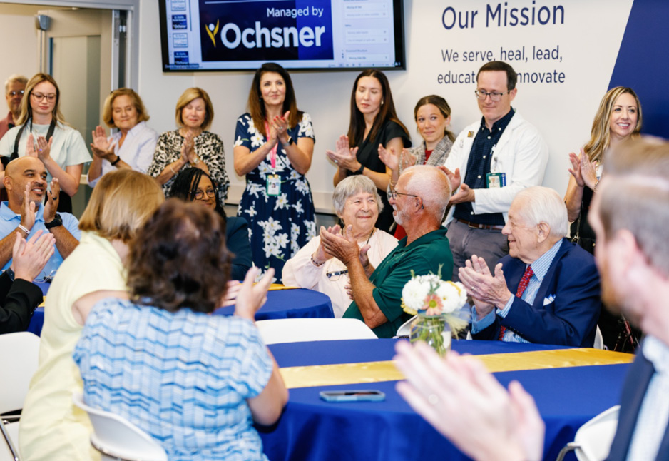 A group of people, some seated and some standing, applaud during an indoor event at an Ochsner-managed facility. A wall sign in the background displays the organization's mission statement.